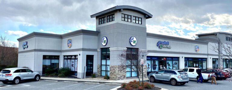 A strip mall with a gray exterior features several businesses, including a vitamin shop and a fitness center. Cars are parked in front of the stores, which are surrounded by a cloudy sky and trees.