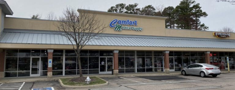 A strip mall with two visible storefronts: Comfort Medical Supply and Firehouse Subs. The parking lot has a few cars, and bare trees are visible next to the sidewalk under a cloudy sky.