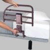 A person installs a Freedom Click Extendable Bed Rail, featuring a brown three-bar design, on the side of a mattress with white sheets and a pillow. The bed rail is secured to the bed frame.