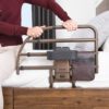 A person in a burgundy sweater and beige pants sits on a bed, holding the Freedom Click Extendable Bed Rail with an attached storage pouch and sturdy frame.