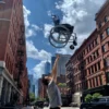 A person is playfully tossing a Feather Light 18 Wheelchair into the air on a sunny city street, surrounded by red brick buildings and a sky dotted with scattered clouds.