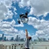 Near a waterfront with skyscrapers in the background and a partly cloudy sky, someone tosses a Feather Light 18 Wheelchair into the air.