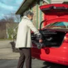 An older adult in a beige coat places a folded Feather Light 18 Wheelchair into the trunk of a red car parked in a driveway, with a house and leafless trees in the background.