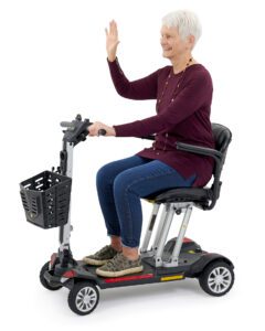 A senior with short white hair smiles and waves while seated on a Golden Buzzaround Carry On mobility scooter featuring a black seat, handlebars, and front basket. They wear a maroon top and blue jeans.