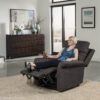 A woman smiles while relaxing in the dark Pride VivaLift!® Urbana 2 Model PLR-965 recliner, feet up and drink in hand. The modern room features a large window, artwork, a wooden dresser, and a beige rug.