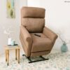 A Pride VivaLift!® Urbana 2 Collection Model PLR-965 tan lift chair stands upright in a cozy room. A small side table with a white teapot and flowers is on the left. A round patterned rug and a decorative vase with branches adorn the floor. A remote rests on the chair.