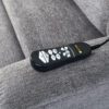 A Golden Technologies®️ Comforter with MaxiComfort PR 535 remote control, featuring buttons for up, down, massage, and heat functions, is on the gray armrest of a sofa.