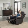 A person lounges in a Pride VivaLift!® Urbana 2 Collection Model PLR-965 recliner, drink in hand, dressed casually. The modern living room includes a sideboard with decor, framed art on the walls, and a large window bathing the space in natural light.