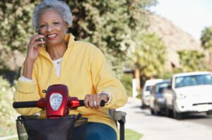 An elderly woman in a yellow jacket smiles while talking on a mobile phone. She is sitting on a mobility scooter on a sunny street lined with parked cars and trees.
