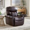 A Golden Technologies®️ Cloud+ Power Lift Recliner PR 511 in dark brown leather sits on a light rug in a living room. Nearby, a modern wooden side table holds a cup and book, while in the background stands a wooden sideboard adorned with plants and decor. Natural light fills the space.
