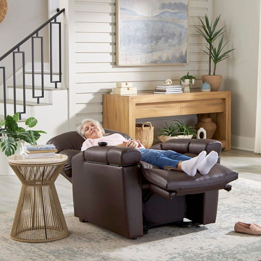 An elderly person is comfortably reclining on a Golden Technologies®️ Cloud+ Power Lift Recliner PR 511 in a cozy living room. Nearby, a small round table with a plant sits alongside a wooden console adorned with decor. Stairs featuring a black railing are visible on the left.