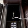 An open compartment in the Golden Technologies®️ Cloud+ Power Lift Recliner PR 511s armrest reveals a USB port with a charging cable connected, along with a visible remote control. The recliner features brown leather upholstery.