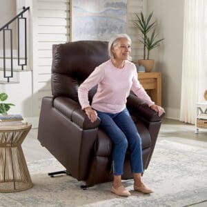 An elderly woman in a pink sweater and jeans uses the Golden Technologies®️ Cloud+ Power Lift Recliner PR 511 to assist standing up in a cozy, well-lit living room, accented by a potted plant and light-colored rug.