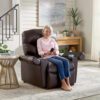 An elderly woman with gray hair sits in a Golden Technologies®️ Cloud+ Power Lift Recliner PR 511, using a remote control. She wears a pink sweater and blue jeans, in a bright room with a side table, painting, and houseplants.