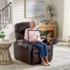 An elderly woman relaxes in a Golden Technologies®️ Cloud+ Power Lift Recliner PR 511, reading a book. She wears glasses, a light pink sweater, and blue pants. Nearby are a table with a cold drink, wooden console, and houseplants. The room is bright with natural light.