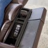 The Golden Technologies®️ Cloud+ Power Lift Recliner PR 511 in brown leather features an open armrest compartment with storage for a remote control and phone. The armrest has two sections, including one with a textured surface. A persons leg is partially visible on the recliner.