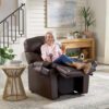 An older woman smiles while seated on a Golden Technologies®️ Cloud+ Power Lift Recliner PR 511, holding a TV remote and a drink. A rustic wooden table with plants adds to the cozy living room, featuring a staircase and landscape painting in the background.
