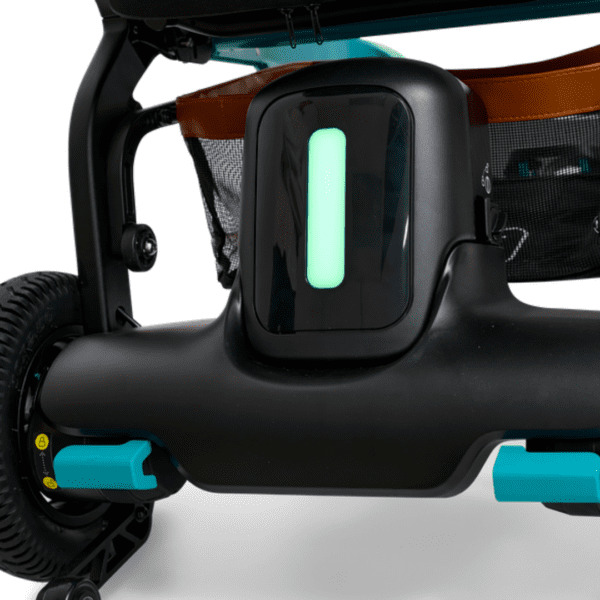 A close-up of a modern strollers wheel and base features a glowing green rectangular light on a sleek black component. In the background, theres a brown storage basket with a black mesh panel, and the tire boasts thick tread and teal accents, similar to the Golden Ally Power Wheelchair.