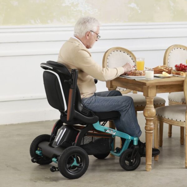 An elderly man, dressed in a beige sweater and blue jeans, sits in a Golden Ally Power Wheelchair at the dining table, enjoying breakfast with assorted dishes and a glass of orange juice as he basks in the bright rooms warmth.