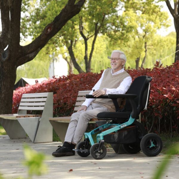 A smiling elderly man with white hair and glasses relaxes in his Golden Ally Power Wheelchair, taking in the lush greenery and red bushes along the path. A sleek bench sits beneath towering trees, enhancing this sunny day scene.