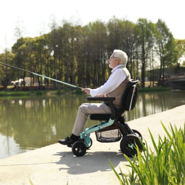 An elderly man fishes by a lake seated in a Golden Ally Power Wheelchair, wearing a white shirt and beige vest. Trees and greenery frame the serene scene under a clear sky.