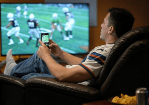 A man sitting in recliner chair watching a football game. He's got his phone in his hands and snacks by his side.